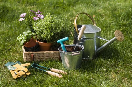 A neatly maintained front garden with a lush green lawn area, edged by a flower bed containing potted plants and flowering shrubs in full bloom, situated in a residential area in Wembley. The garden features a well-kept grass surface with visible mowing lines, surrounded by natural soil borders and a paved pathway partially visible in the background. Gardening tools such as a pair of yellow gardening gloves, a small trowel, and pruning shears are laid out on the grass, alongside a metal watering can equipped with a long spout, and a silver bucket with a handle, indicating recent gardening activity. The setting suggests a typical outdoor space prepared for routine garden care and hedge trimming, reflecting professional gardening and landscaping services available in Wembley. The lighting indicates daytime with natural sunlight illuminating the scene, showcasing a tidy and inviting outdoor environment suitable for various garden maintenance tasks.