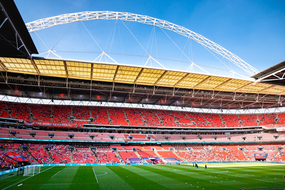 An indoor stadium with a partially open, curved roof structure featuring white support beams and a yellow underside canopy. The seating areas are predominantly red and are arranged in multiple tiers s
