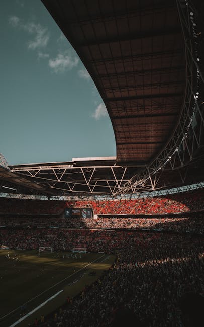 An indoor stadium with a partially open, curved roof structure featuring white support beams and a yellow underside canopy. The seating areas are predominantly red and are arranged in multiple tiers s